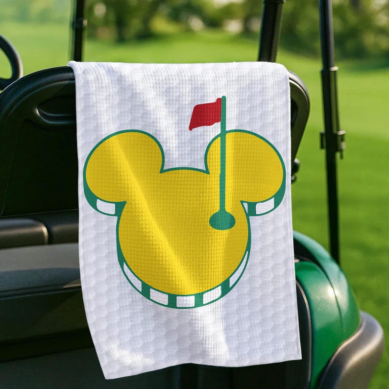 May include: White golf towel with a yellow Mickey Mouse-shaped golf course logo and a red flag. The towel is hanging on a golf cart. The logo has a green outline and a green golf pin.