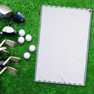 May include: A golf scene featuring golf clubs, golf balls, and a white golf towel with a green border. The items are arranged on a vibrant green grass surface, creating a clean and organized display.