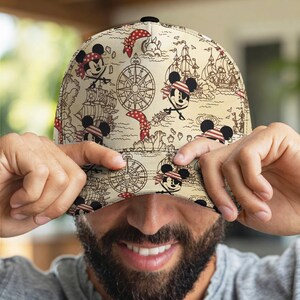 May include: A beige baseball cap featuring a pirate-themed design with Mickey Mouse illustrations, compasses, and pirate ships. The cap has a black button on top and a curved brim. The design includes red and white accents, creating a playful and adventurous aesthetic.