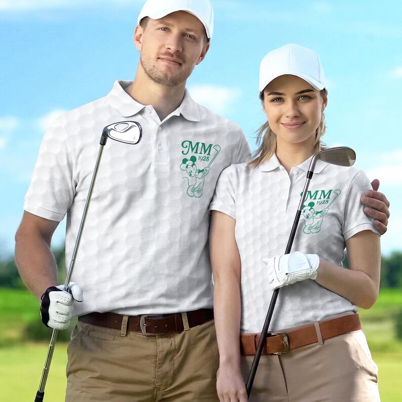 K&ouml;nnte beinhalten: Zwei Personen tragen wei&szlig;e Golf-Poloshirts mit einem Golfballmuster und einer gr&uuml;nen Grafik eines Zeichentrickcharakters und den Buchstaben "MM 1928". Sie halten Golfschl&auml;ger und tragen wei&szlig;e Kappen.