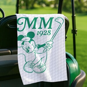 K&ouml;nnte beinhalten: Wei&szlig;es Golfhandtuch mit gr&uuml;ner Grafik von Mickey Mouse beim Golfspielen. Das Handtuch zeigt die Initialen "MM" und das Jahr "1928" in Gr&uuml;n. Das Handtuch h&auml;ngt an einem Golfwagen.