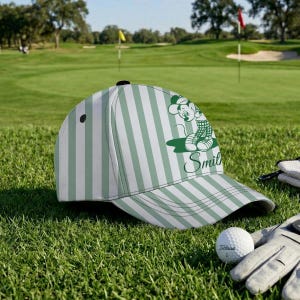 May include: A white and green striped baseball cap with a cartoon character design and the name "Smit" in green script. The cap is on a golf course, next to a golf ball and glove.