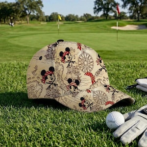 May include: Tan baseball cap featuring a pirate-themed design with Mickey Mouse illustrations, ships, compasses, and anchors. The cap is on a golf course, next to a golf ball and glove.