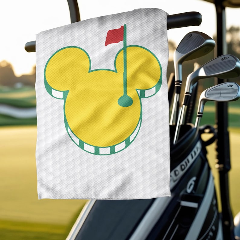May include: White golf towel featuring a yellow Mickey Mouse head design with green and red accents. The towel has a golf flag graphic and is hanging on a golf cart. Golf clubs are in the background.