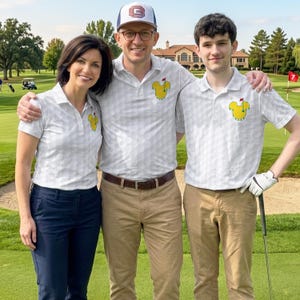May include: Three people pose on a golf course, wearing matching white polo shirts with a yellow cartoon character design. The shirts have a subtle geometric pattern. One person wears a baseball cap with a logo. The background includes a golf course and a building.