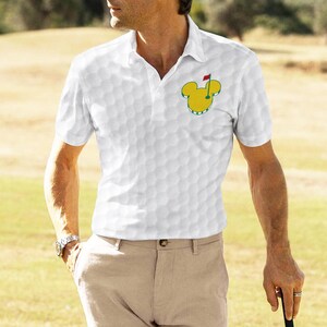 May include: White polo shirt with a golf ball pattern and a Mickey Mouse golf logo. The shirt has a classic collar and short sleeves. The golf logo is yellow and green with a red flag.