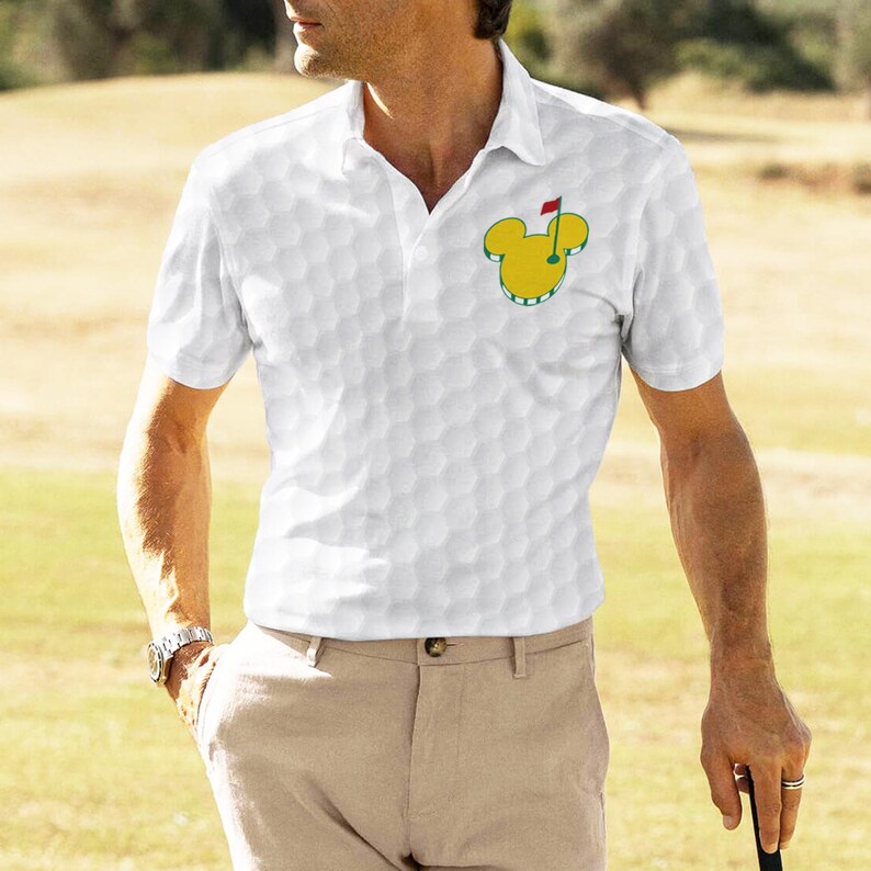 May include: White polo shirt with a golf ball pattern and a Mickey Mouse golf logo. The shirt has a classic collar and short sleeves. The golf logo is yellow and green with a red flag.