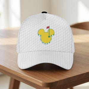 May include: White baseball cap with a golf ball texture. The cap features a yellow Mickey Mouse-shaped golf course logo with a red flag. The cap is sitting on a wooden table.