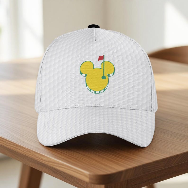 May include: White baseball cap with a golf ball texture. The cap features a yellow Mickey Mouse-shaped golf course logo with a red flag. The cap is sitting on a wooden table.