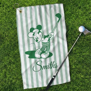 May include: A golf towel with a green and white striped pattern. The towel features a green cartoon image of Mickey Mouse swinging a golf club. The name "Smiths" is printed in green script below the cartoon. A golf club and ball are also in the image.
