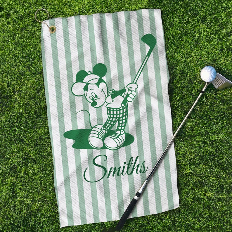 May include: A golf towel with a green and white striped pattern. The towel features a green cartoon image of Mickey Mouse swinging a golf club. The name "Smiths" is printed in green script below the cartoon. A golf club and ball are also in the image.