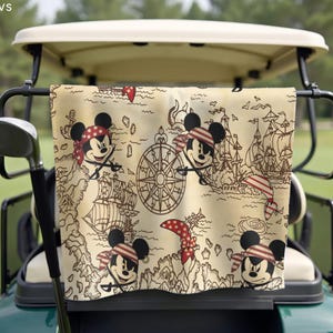 May include: A beige golf towel featuring a pirate-themed map design with illustrations of Mickey Mouse in a red and white striped bandana. The towel also includes images of pirate ships, a compass, and a red polka-dot bandana.