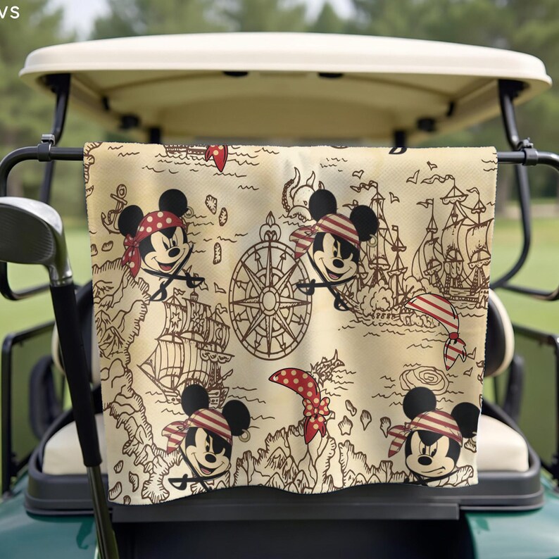 May include: A beige golf towel featuring a pirate-themed map design with illustrations of Mickey Mouse in a red and white striped bandana. The towel also includes images of pirate ships, a compass, and a red polka-dot bandana.