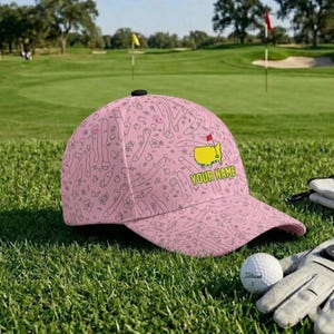 Może przedstawiać: Różowa czapka z daszkiem z wzorem pola golfowego i napisem "YOUR NAME". Czapka posiada żółte logo pola golfowego z czerwoną flagą. Piłka golfowa i rękawica znajdują się na pierwszym planie.