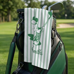 May include: A green and white striped golf towel with a cartoon character graphic of a mouse playing golf. The towel is hanging on a golf bag with golf clubs. The name "Smiths" is printed on the towel.