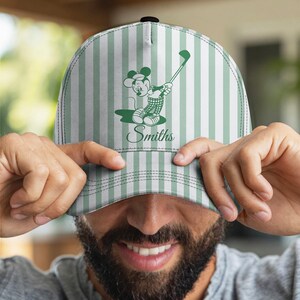 May include: A green and white striped baseball cap featuring a cartoon mouse playing golf. The cap has the name "Smiths" in script. The cap is being held up by a person.