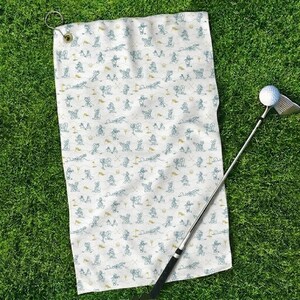 Może przedstawiać: Biały ręcznik golfowy z powtarzającym się wzorem kresk&oacute;wkowych myszy grających w golfa w kolorze niebieskim i złotym. Kij golfowy i piłka znajdują się po prawej stronie, na zielonej trawie. Ręcznik ma metalową przelotkę do zawieszania.