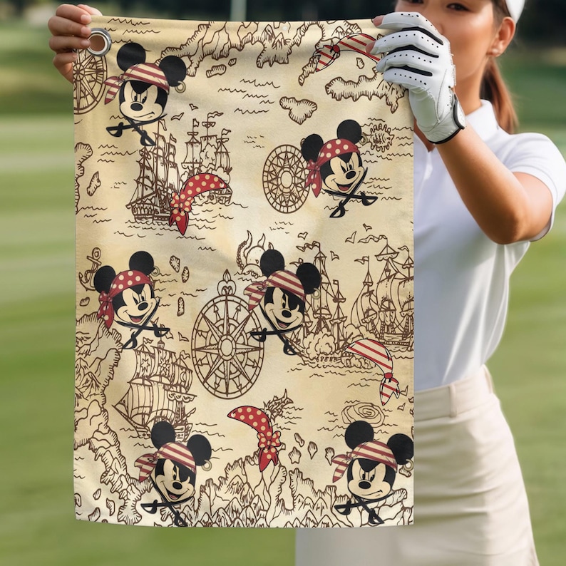 May include: Golf towel featuring a pirate-themed design with Mickey Mouse characters, pirate ships, and treasure map elements. The towel has a beige background with red, black, and white accents. The design includes compasses and crossed swords.