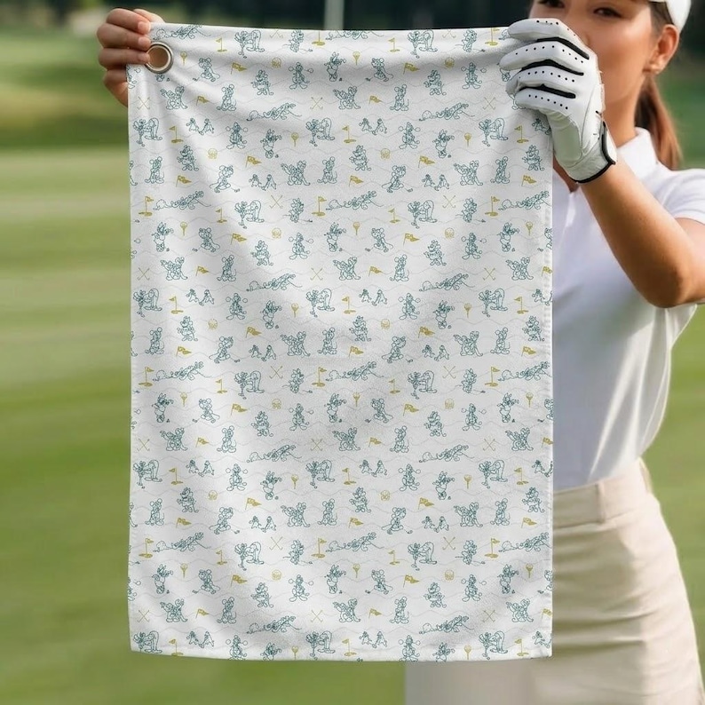 Może przedstawiać: Biały ręcznik golfowy z powtarzającym się wzorem kresk&oacute;wkowych golfist&oacute;w, piłek golfowych, flag i kij&oacute;w w kolorze morskim i złotym. Ręcznik ma metalową przelotkę do zawieszania.