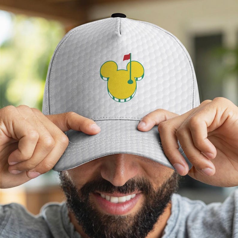 May include: White golf cap featuring a yellow Mickey Mouse-inspired design with a golf flag. The cap has a textured pattern and a black button on top. The design is outlined in green and includes a red flag.