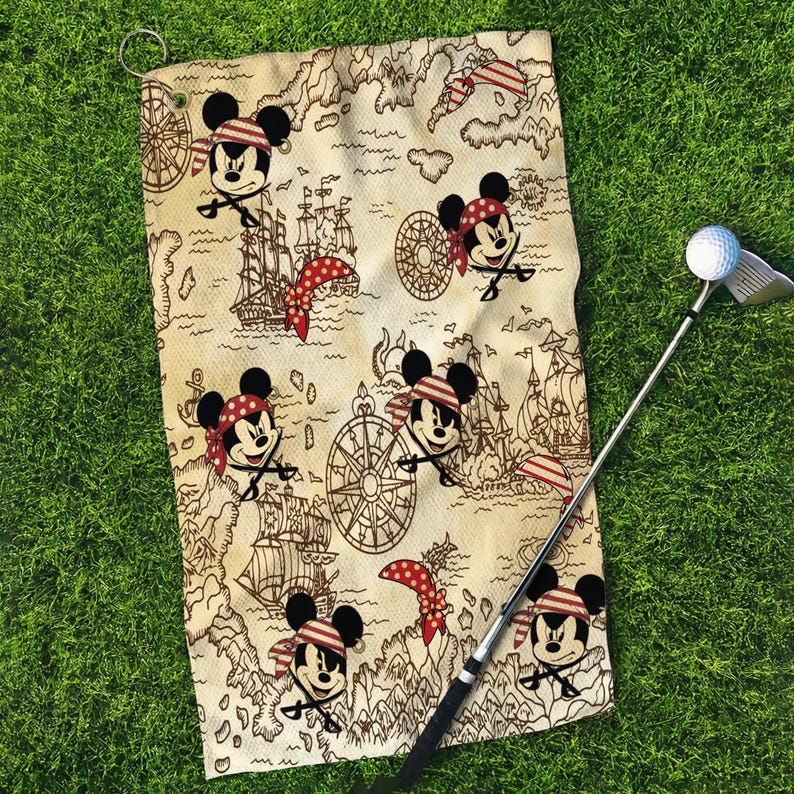 May include: Golf towel with a pirate-themed design featuring Mickey Mouse characters, pirate ships, and compasses on a map-like background. A golf club and ball are visible on the green grass.