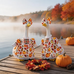 Può includere: Due oche vestite con abiti e fiocchi decorati con zucche e foglie autunnali sono su un molo di legno. Anche zucche e una ghirlanda di foglie sono sul molo, con un lago e alberi autunnali sullo sfondo.