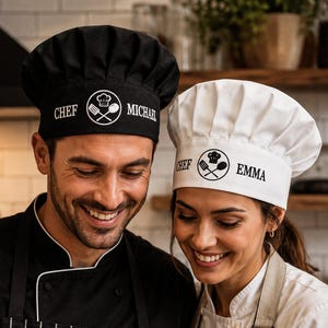 May include: A black chef hat with the text CHEF MICHAEL and a white graphic of a chef hat, spatula, and spoon, alongside a white chef hat with the text CHEF EMMA and a black graphic of a chef hat, spatula, and spoon, both featuring pleated crowns.