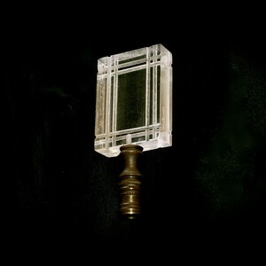 May include: A clear, rectangular bottle stopper with a geometric design. The stopper features a square top with a grid pattern and a bronze-colored base. The design is reminiscent of an architectural window.