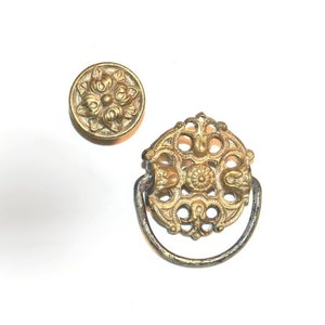 May include: Two gold-toned metal cabinet knobs with ornate floral designs. One knob is round and the other is a ring pull.