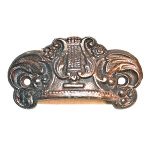 May include: A vintage, ornate, metal drawer pull with a lyre design in the center. The pull is a dark bronze color and has a decorative, scroll-like design.