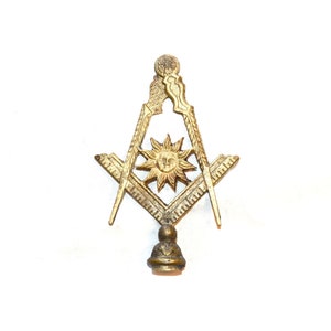 May include: A gold-colored metal Masonic symbol with a compass and square, a sun with a face in the center, and a decorative base.
