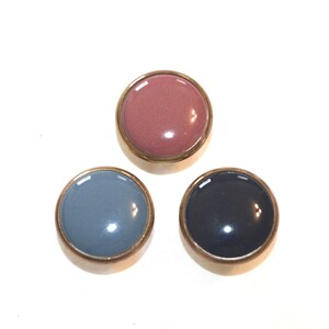 May include: Three round buttons with a metallic gold rim. The buttons have glossy, solid-color centers in dusty rose, slate blue, and navy blue. The buttons are arranged in a triangular pattern against a white background.