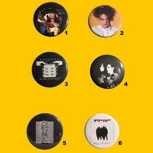Può includere: Una collezione di sei spille rotonde con vari disegni. Le spille presentano immagini di musicisti e loghi di band, tra cui Kate Bush, The Cure, Bauhaus e Joy Division. Le spille sono su uno sfondo giallo.