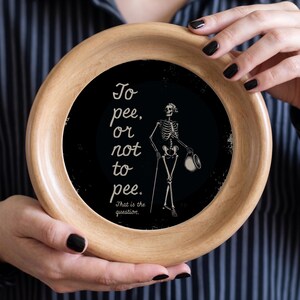 May include: A circular wooden decorative plate with a black centre. It features a skeleton illustration holding a cane and hat, with the text "To pee, or not to pee. That is the question."