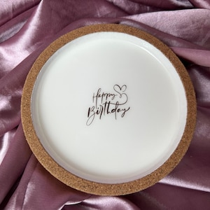 Könnte beinhalten: Ein runder weißer Keramik-Tablett mit einem Korksockel. Auf dem Tablett steht in schwarzer Schrift "Happy Birthday". Das Tablett befindet sich vor einem Hintergrund aus glänzendem, malvenfarbenem Stoff.