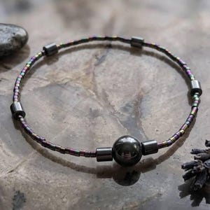 Op de afbeelding: Een kralenarmband met een grote, donkergrijze centrale kraal en kleinere cilindrische kralen. De draad van de armband heeft iriserende kleuren. De armband wordt getoond op een getextureerd grijs oppervlak met donkergrijze stenen en een takje lavendel.