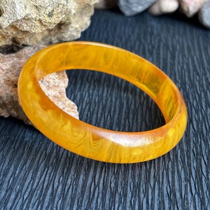 Op de afbeelding: Een gladde, cirkelvormige armband van doorschijnende, gemarmerde, amberkleurige hars. De armband heeft een gepolijste, glanzende afwerking met wervelende patronen van lichtere en donkerdere geeltinten door de gehele massieve, ronde band.