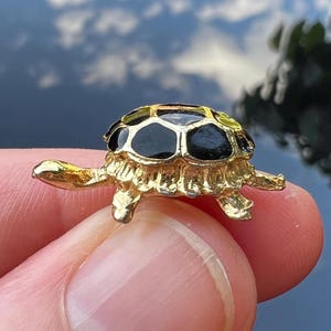以下が含まれることがあります： 黒と緑の甲羅を持つ金色のカメの置物。カメは小さく、足と頭が伸びています。甲羅には幾何学模様があります。置物は2本の指の間に挟まれています。