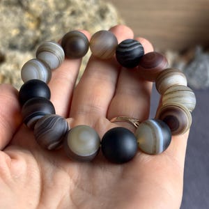 Puede incluir: Una pulsera de cuentas compuesta por piedras redondas con acabado mate en varios tonos de gris, negro, marrón y crema. Las cuentas muestran patrones naturales de bandas, algunas son de color negro sólido o gris translúcido, en un hilo elástico.
