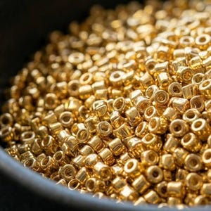 Könnte beinhalten: Nahaufnahme eines Haufens kleiner, glänzender, goldfarbener Perlen. Die Perlen sind zylindrisch mit einem Loch in der Mitte. Die Perlen sind dicht gepackt und füllen den Rahmen aus. Diese Perlen werden wahrscheinlich für die Schmuckherstellung oder andere Bastelarbeiten verwendet.