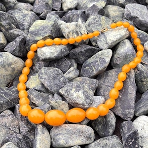 Può includere: Una collana vintage color ambra con perline ovali. La collana presenta una chiusura color oro ed è esposta su un letto di rocce grigie e nere. Le perline variano di dimensioni, con perline ovali più grandi al centro.