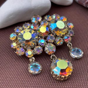 Peut inclure: Broche vintage ornée de strass irisés multicolores. La broche présente des grappes de pierres rondes et ovales dans les tons jaune, bleu et violet. Deux pierres pendantes sont suspendues en bas. Monture en métal argenté.