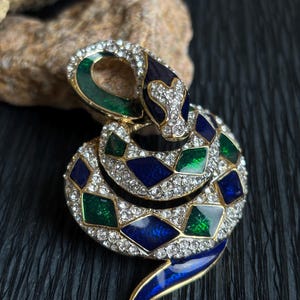 Puede incluir: Un broche de serpiente enroscada de metal dorado, con incrustaciones de esmalte en forma de diamante en azul oscuro y verde esmeralda, y pedrería transparente engastada en pavé que cubre el cuerpo y la cabeza de la figura serpentina.