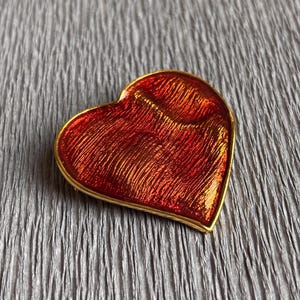 Puede incluir: Un broche en forma de corazón con una incrustación de esmalte naranja rojizo metálico vibrante y una superficie texturizada con ondas. El corazón está enmarcado por un borde fino de metal dorado pulido que sigue el contorno curvo.