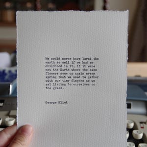 May include: A rectangular piece of white paper with deckled edges featuring black typewriter-style text that reads: We could never have loved the earth so well if we had no childhood in it, if it were not the Earth where the same flowers come up again every spring that we used to gather with our tiny fingers as we sat lisping to ourselves on the grass. George Eliot.