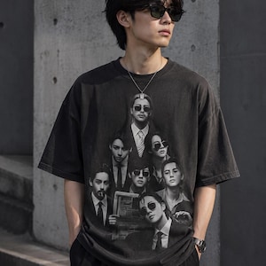 以下が含まれることがあります： 黒のグラフィックTシャツ。グレーの階調でスーツとサングラスをかけた人々の画像がプリントされています。シャツはヴィンテージ風で色あせた外観で、黒いパンツと合わせています。全体的なスタイルはカジュアルでモダンです。