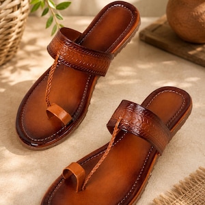 May include: A pair of brown leather sandals featuring a wide, embossed strap across the instep and a thin, braided leather cord connecting to a toe loop. The sandals have a smooth, polished finish with darker shading along the edges of the soles.