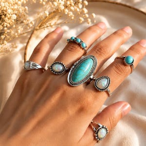 Könnte beinhalten: Eine Sammlung von sieben silbernen Ringen an den Fingern, besetzt mit verschiedenen Steinen, darunter ein großer ovaler türkisfarbener Cabochon, mehrere kleinere irisierende opalartige Steine und kleine runde türkisfarbene Perlen in Silber.