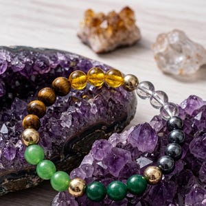 Könnte beinhalten: Ein mehrfarbiges Perlenarmband mit grünen, goldenen, braunen, gelben, klaren und schwarzen Perlen. Das Armband wird auf einem Bett aus lila Amethystkristallen präsentiert, mit weiteren Kristallen im Hintergrund.