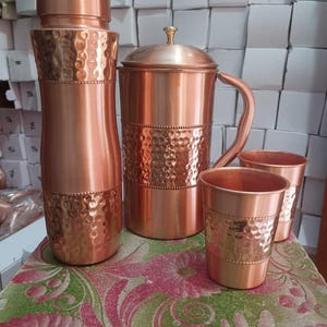 May include: A set of copper drinkware consisting of a tall water bottle, a pitcher with a lid and handle, and two tumblers. Each item features a polished finish with a central band of hammered texture and a decorative beaded border detail.